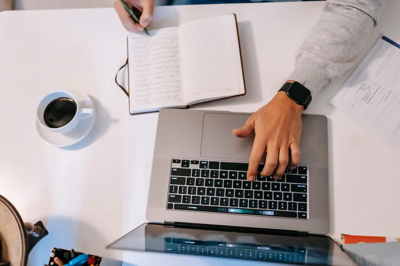 Person typing on a laptop while making notes in a journal with a cup of coffee nearby
