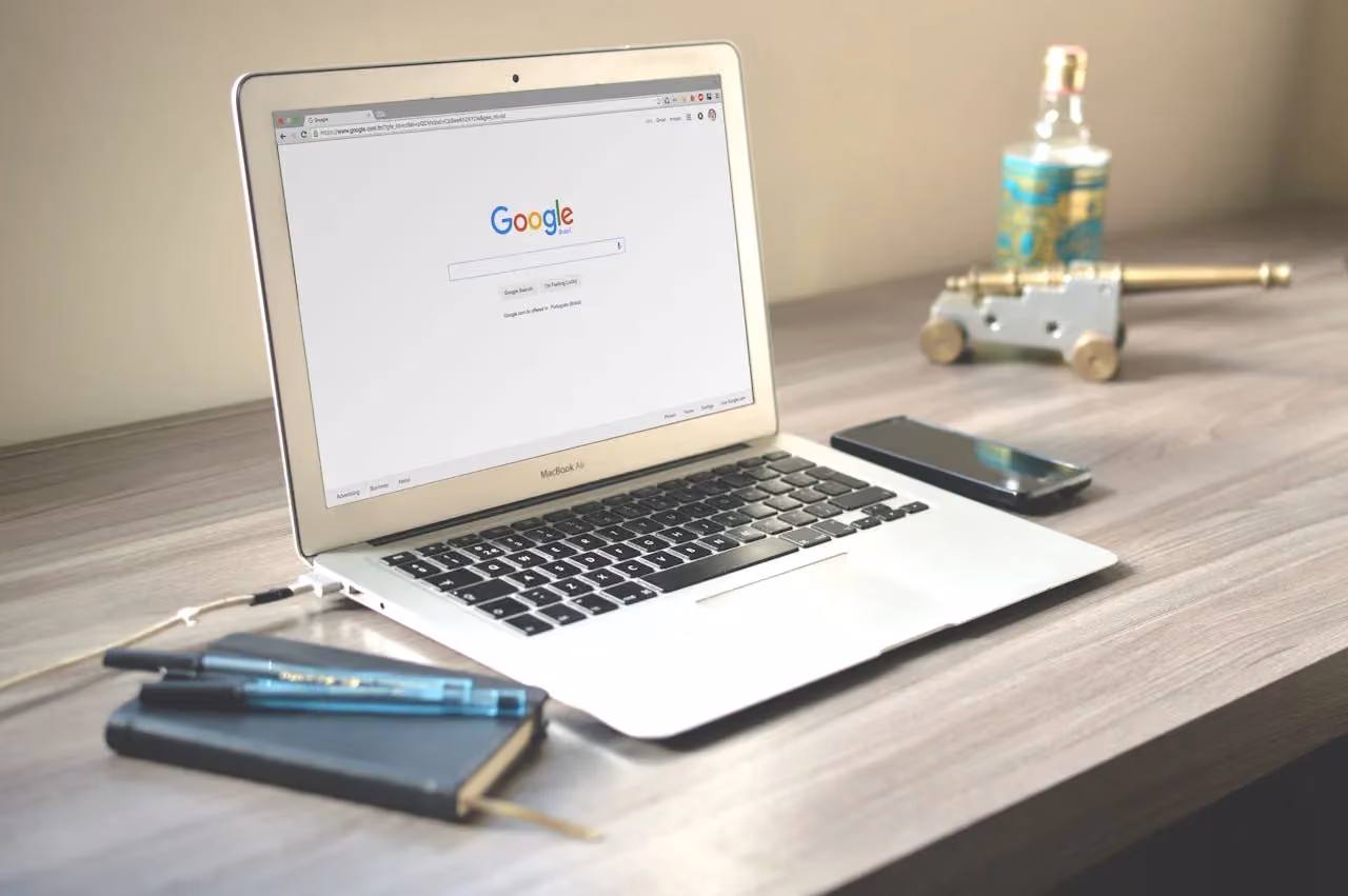MacBook Air on a desk displaying the Google search homepage with a notebook and phone alongside