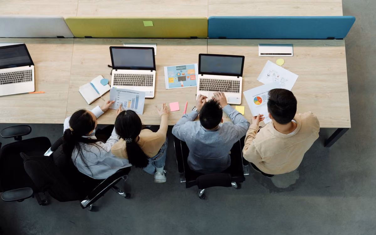Four colleagues working on laptops at a shared office desk, seen from above