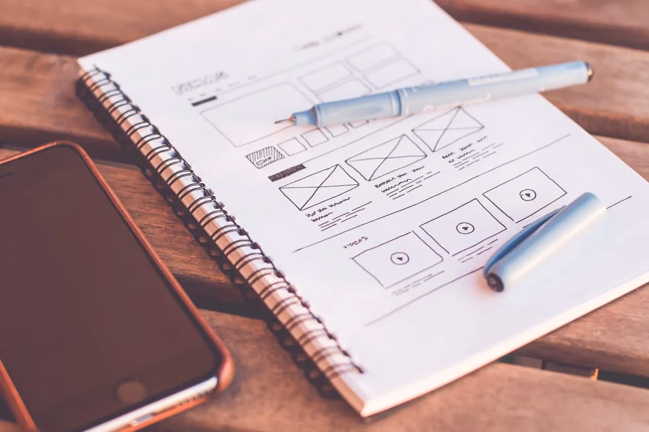 Notebook with wireframe sketches beside a smartphone on a wooden desk, planning a website layout